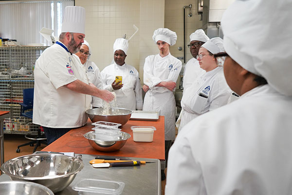Culinary arts students watch an instructor demonstrate professional cooking techniques in an STLCC kitchen classroom.