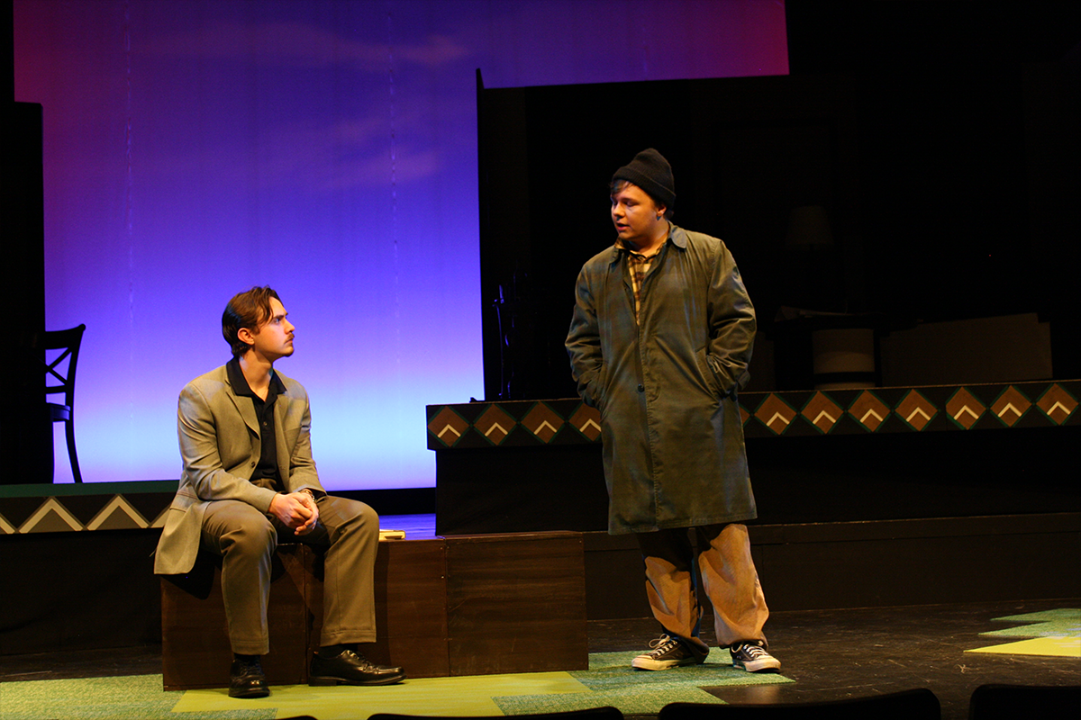 Man in suit sitting on box looking at standing man in beanie and overcoat