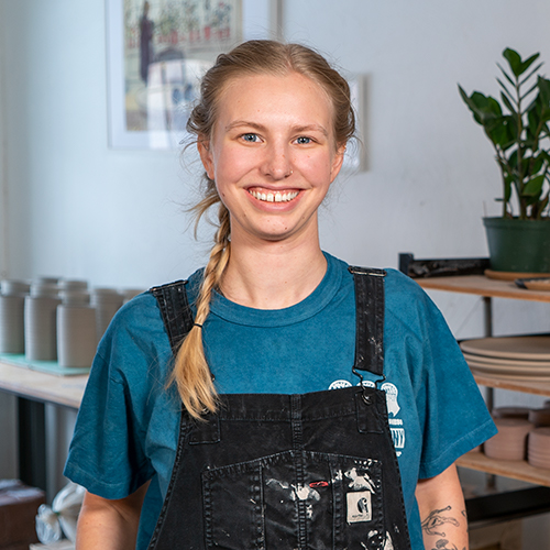 molly svoboda smiles at the camera with a table and shelves behind her full of pottery