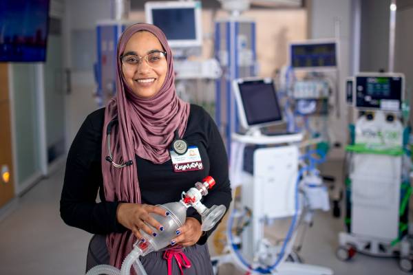 Syeeda Ali stands in front of a hospital room full of respiratory care equipment